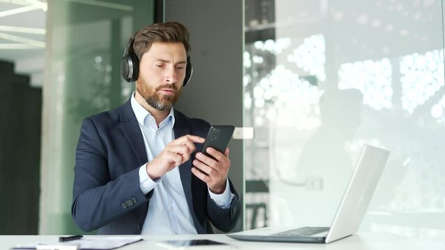 Handsome businessman with headphones listening to relaxing music sitting in business office. Happy entrepreneur turns on playlist on smartphone and enjoys the sounds of relaxation and noise of nature