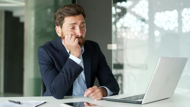 Overworked businessman in formal suit is bored sitting at workplace at desk in business office. Exhausted worker is tired from working with a laptop. A lazy worker procrastinates, gets distracted