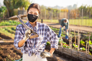 Portrait of confident female gardener in protective face mask standing with shovel during spring...