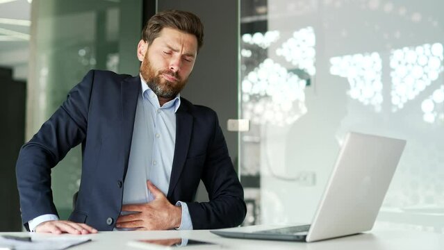 Businessman in a formal suit feels stomach pain while working on a laptop while sitting at workplace in business office. Sick worker has heartburn, gastritis or poisoning. Male suffers from spasms