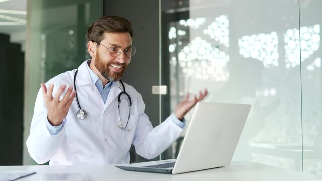 Excited happy doctor in white coat received great news on laptop sitting in modern hospital clinic. Joyful medical worker physician reads a pleasant message, celebrates success, rejoices at a surprise