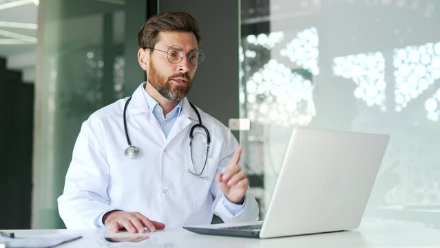 Confident doctor talking on video call using laptop in hospital clinic. Male medical worker physician communicates remotely at a conference in the office or has an online consultation with a patient