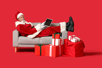 Santa Claus with laptop and gift boxes lying on sofa against red background