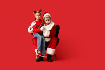 Santa Claus and cute little girl sitting on armchair against red background