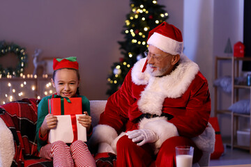 Santa Claus and cute little elf with gift boxes sitting on sofa in decorated room for Christmas at night