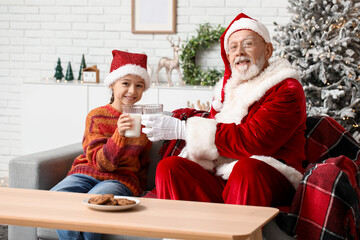 Fototapeta premium Santa Claus and cute little girl drinking milk with cookies on sofa in decorated room for Christmas