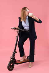 Portrait of young businesswoman with kick scooter drinking coffee on pink background © Pixel-Shot
