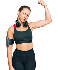 Beautiful blonde woman wearing gym clothes and using headphones strong person showing arm muscle, confident and proud of power