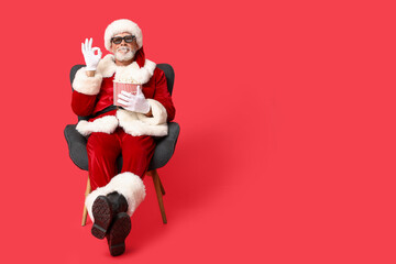 Santa Claus in 3D glasses with bucket of tasty popcorn showing ok gesture on red background