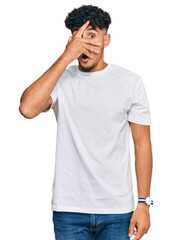 Young arab man wearing casual white t shirt peeking in shock covering face and eyes with hand, looking through fingers with embarrassed expression.