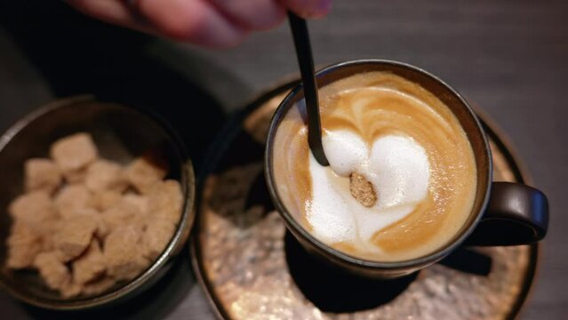Woman stirring brown sugar into a cappuccino cup of coffee with white heart