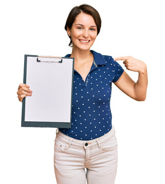 Young brunette woman with short hair holding clipboard with blank space pointing finger to one self smiling happy and proud
