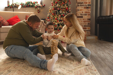 Happy family are sitting on floor near Xmas tree playing board game with wooden tower together at...
