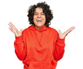 Young hispanic woman with curly hair wearing casual sweatshirt celebrating mad and crazy for success with arms raised and closed eyes screaming excited. winner concept