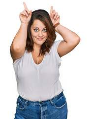 Obraz na płótnie Canvas Young plus size woman wearing casual white t shirt doing funny gesture with finger over head as bull horns
