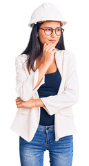 Young beautiful latin girl wearing architect hardhat and glasses with hand on chin thinking about question, pensive expression. smiling with thoughtful face. doubt concept.