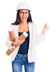 Young beautiful latin girl wearing architect hardhat holding blueprints screaming proud, celebrating victory and success very excited with raised arms