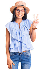 Young beautiful latin girl wearing summer hat and glasses smiling with happy face winking at the...
