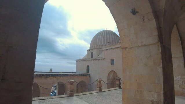 The camera moves towards the dome of Zinciriye Madrasa through the rows of arches. Cut stone workmanship and segmented dome structure can be seen. The sun creates a glare effect.