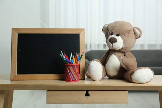 Teddy bear, small blackboard, pencils and pens in holder on wooden table indoors. Space for text
