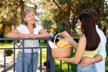 Friendly relationship with neighbours. Young couple with wicker basket of products treating senior woman near fence outdoors