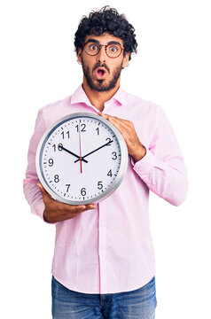 Handsome young man with curly hair and bear holding big clock scared and amazed with open mouth for surprise, disbelief face