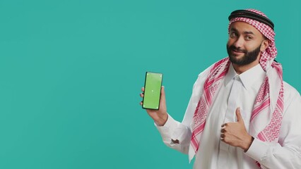 Middle eastern man points at greenscreen display on smartphone, showcasing isolated copyspace template on mobile device screen. Young person in islamic costume shows blank chromakey.