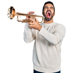 Young hispanic man with beard playing trumpet angry and mad screaming frustrated and furious, shouting with anger looking up.
