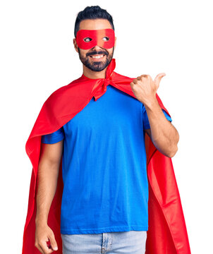 Young hispanic man wearing super hero costume smiling with happy face looking and pointing to the side with thumb up.