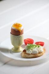 Fresh soft boiled egg in cup and sandwich on white wooden table