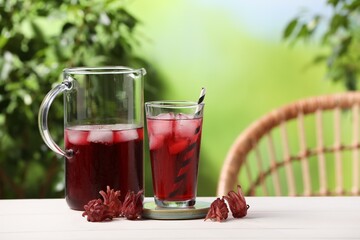 Refreshing hibiscus tea with ice cubes and roselle flowers on white wooden table against blurred green background. Space for text - obrazy, fototapety, plakaty