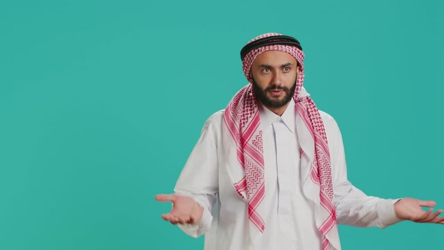 Muslim person being confused and unsure on camera, creating i dont know symbol and acting clueless over blue background. Young man in arab clothing being uncertain and shrugging.