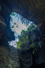 A rectangular opening in a dark, mossy limestone cave frames a bright blue sky with fluffy white clouds in Setigi, Gresik, East Java, Indonesia.