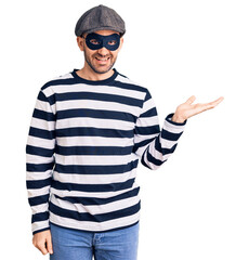 Young handsome man wearing burglar mask smiling cheerful presenting and pointing with palm of hand looking at the camera.