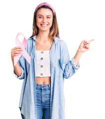 Young beautiful girl holding pink cancer ribbon smiling happy pointing with hand and finger to the side