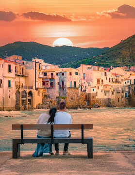 a couple of men and woman sitting on a bench at the waterfront on vacation in Sicily during sunset, a couple visiting the old town of Cefalu Sicilia Italy