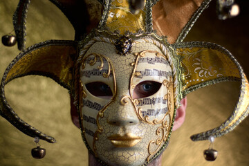 Close up of the face of a young caucasian man with blue eyes wearing a fancy golden Venetian carnival mask on a golden out of focus background