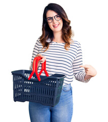 Beautiful young brunette woman holding supermarket shopping basket smiling happy pointing with hand and finger