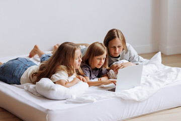 Three beautiful teenage girls are lying on the bed with a laptop and surfing the Internet. Sisters...