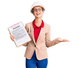 Young beautiful woman wearing architect hardhat holding clipboard with contract document celebrating victory with happy smile and winner expression with raised hands