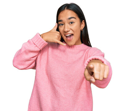 Young asian woman wearing casual winter sweater smiling doing talking on the telephone gesture and pointing to you. call me.