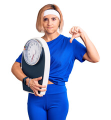 Young blonde woman wearing sportswear holding weighing machine with angry face, negative sign...