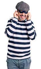 Middle age handsome man wearing burglar mask covering ears with fingers with annoyed expression for the noise of loud music. deaf concept.