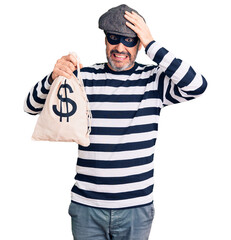 Fototapeta na wymiar Middle age handsome man wearing burglar mask holding money bag stressed and frustrated with hand on head, surprised and angry face