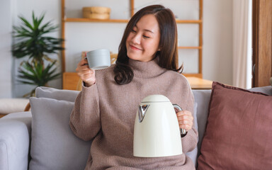 Portrait image of a young woman holding holding a cup and electric kettle at home