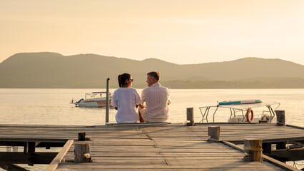 couple sitting on a wooden deck pier in the ocean during sunset in Samaesan Thailand - obrazy, fototapety, plakaty