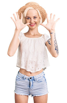 Young blonde woman with tattoo wearing summer hat showing and pointing up with fingers number ten while smiling confident and happy.