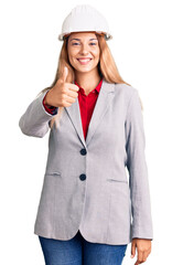 Beautiful young woman wearing architect hardhat doing happy thumbs up gesture with hand. approving expression looking at the camera showing success.