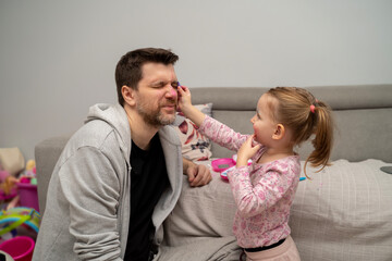 Cute little girl putting on makeup on her father, father making grimace 