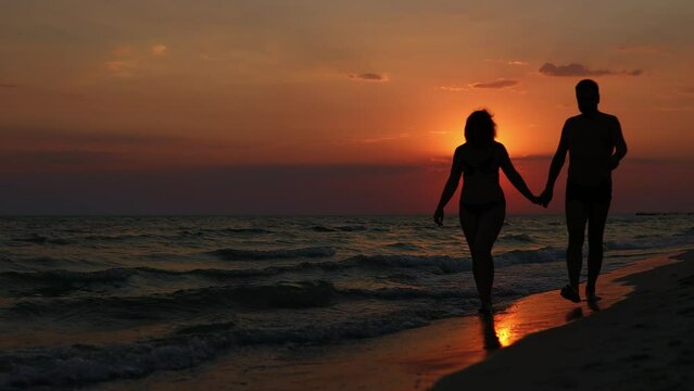 A man and a woman are walking along the seashore holding hands in front of a beautiful sunset. Slow Motion